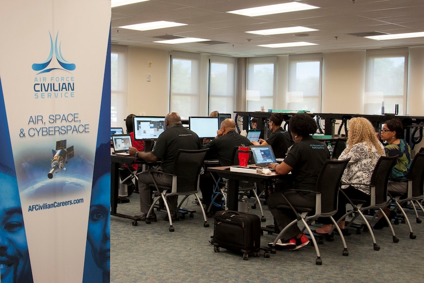 A group of people sit at computer stations in a room. A sign on the side of the room reads, "Air Force Civilian Service."