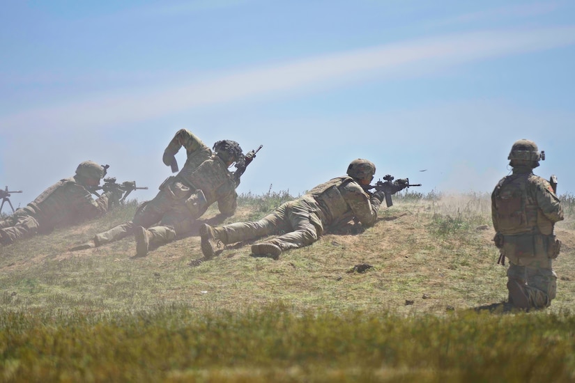 Uniformed service members kneel and lay on an elevated grassy area with their weapons.