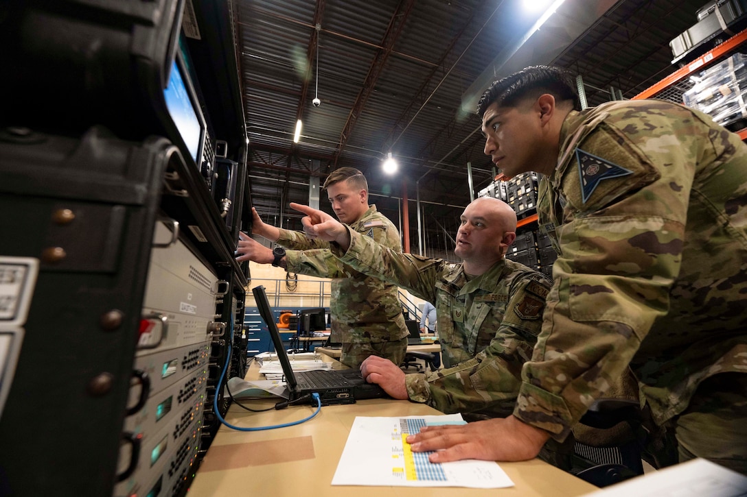 An airman points at a monitor while sitting in between two standing guardians in a warehouse.