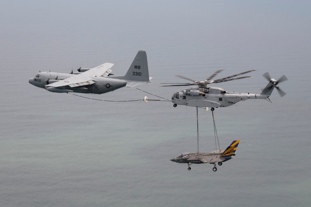 An airborne aircraft connects its boom to a helicopter carrying a small aircraft by sling over a body of water.