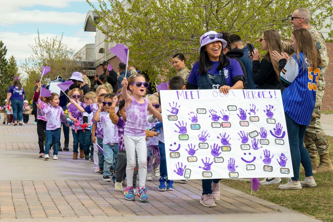 A person carrying a sign of handprints leads a parade of small children wearing purple gear waving flags as service members cheer from the side.