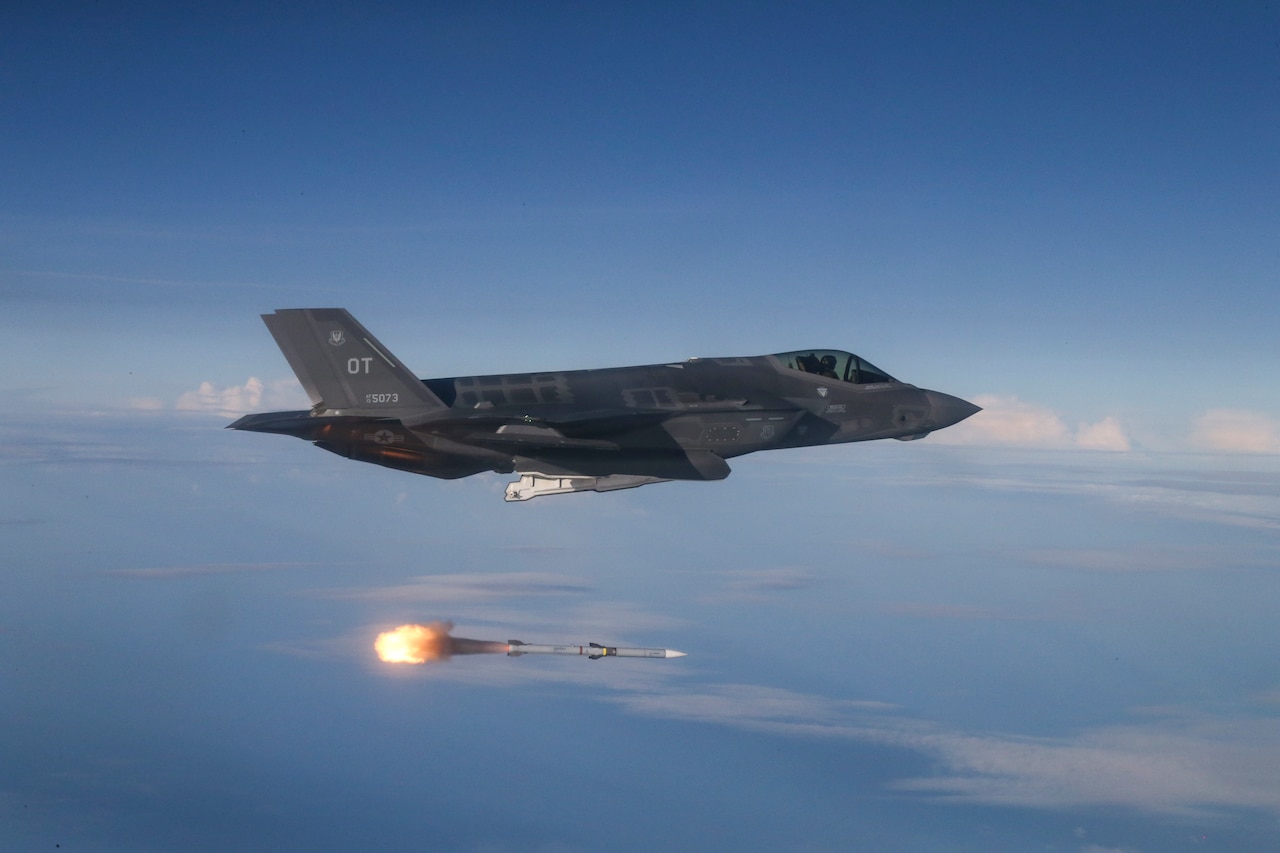 A jet fighter aircraft flying in the sky from left to right. A missile with flames coming out the back is beneath the aircraft, shooting from left to right.