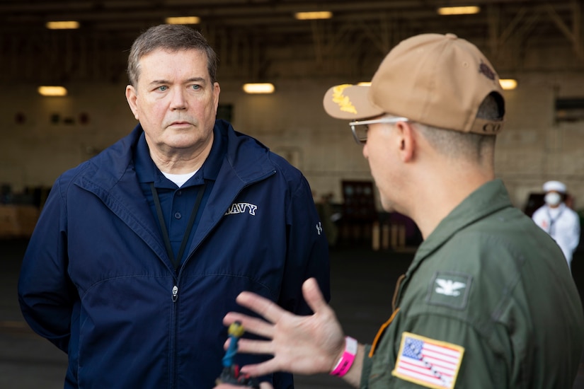 A man in a flight suit and ball cap is in the foreground talking and gesturing with his hands to another man wearing a civilian jacket that says “Navy”.