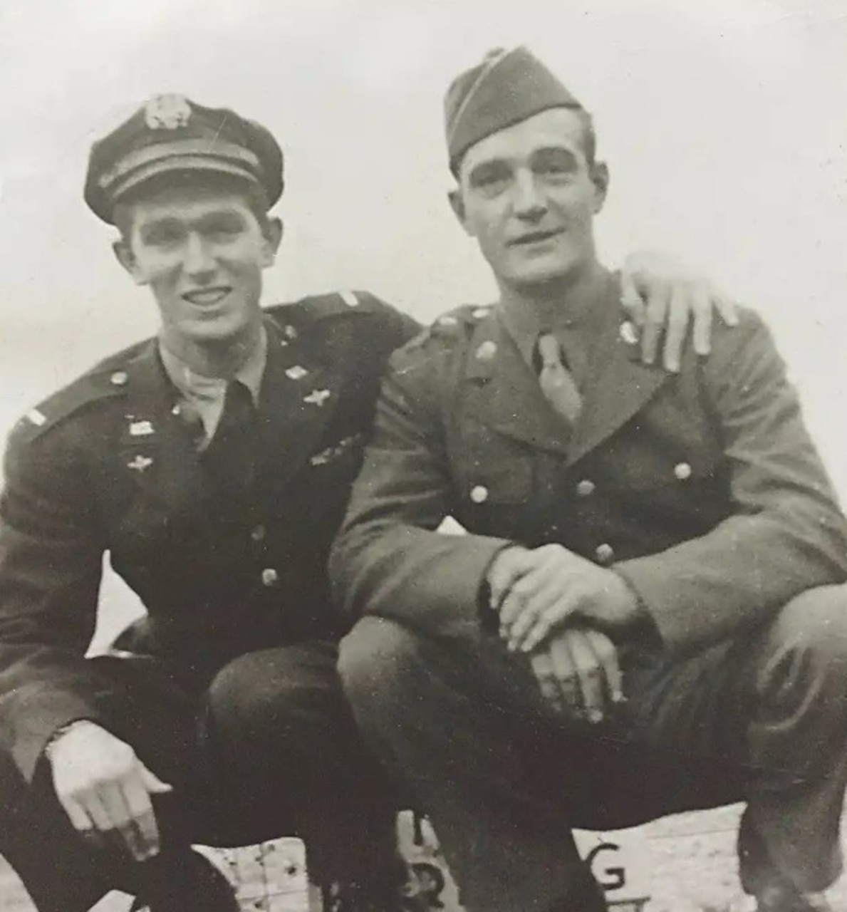 Two men squat down beside each other for a photo. One has his hand on the other’s shoulder.