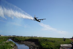 A military C-130J aircraft discharges a pressurized water stream from the rear of the aircraft over a green marshland with a creek affixed against a light blue sky.