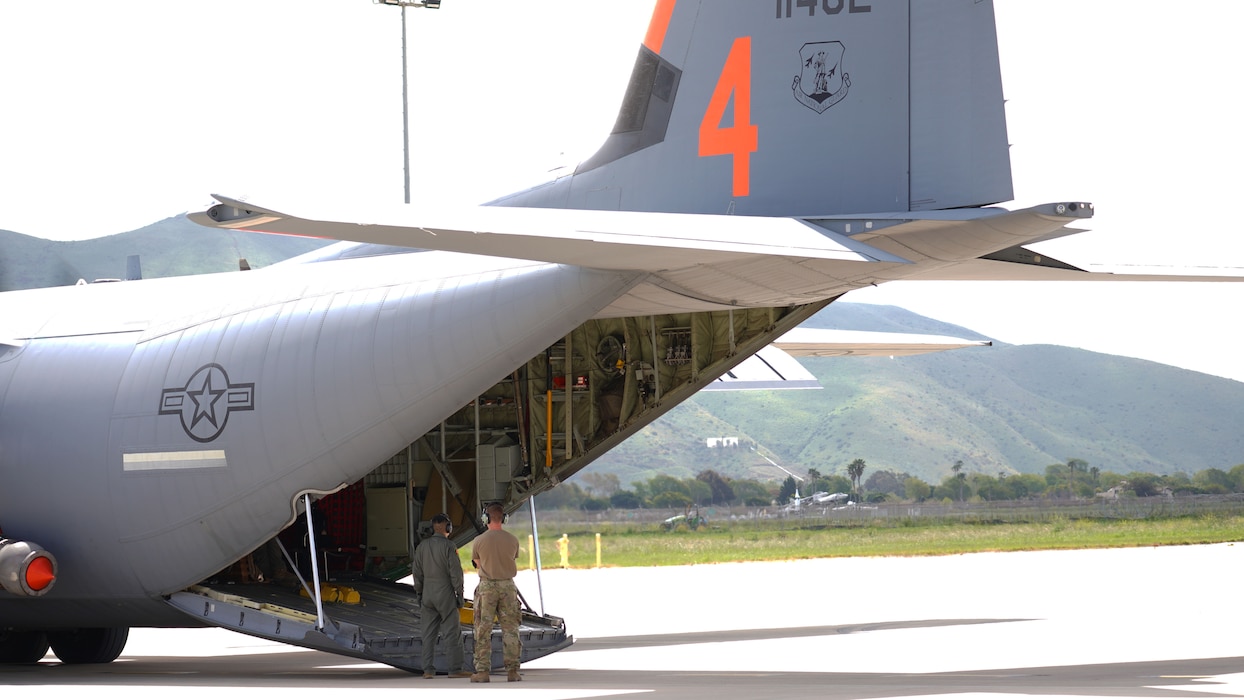 Two military men look into the back of a C-130J with the cargo ramp down on a flightline.