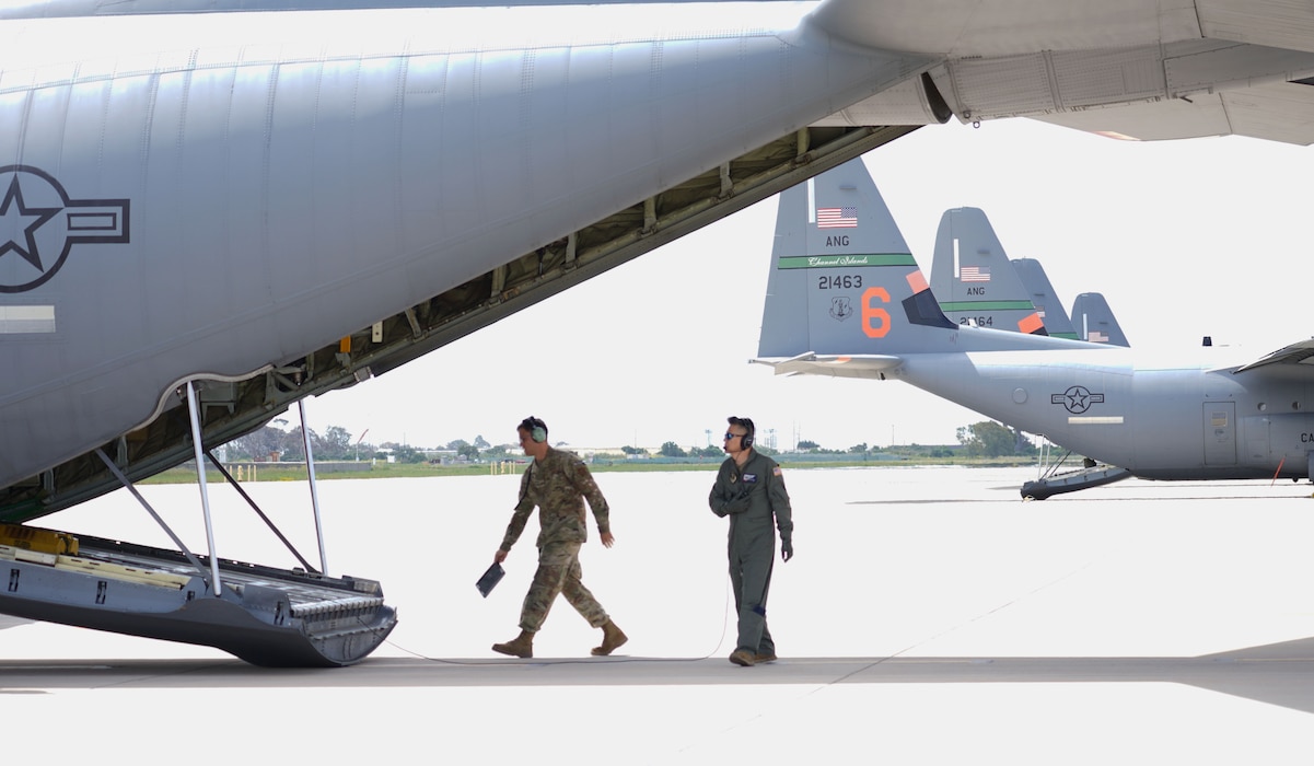 Two military personnel walk towards the back of a lowered ramp of a C-130J on a military flightline.