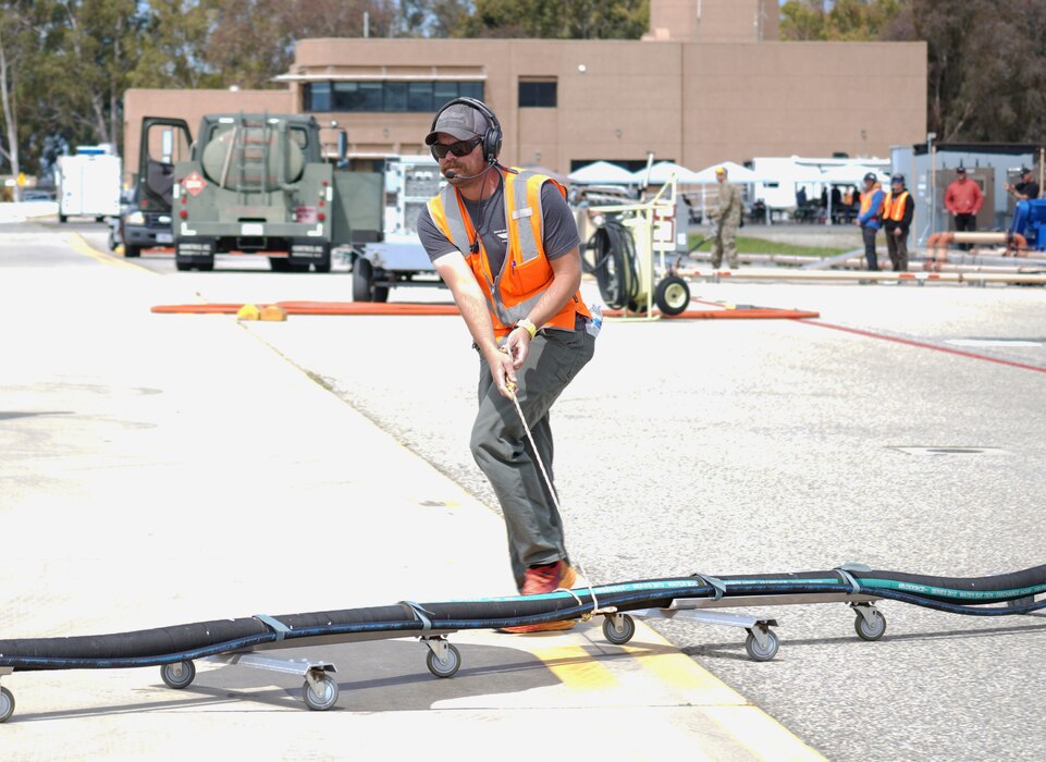 A man pulls a rope attached to a long hose with dolly wheels underneath it to move it across the flight line on a military installation.