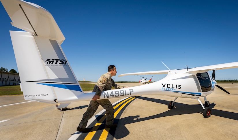 An airman pushes the tail of a small electric aircraft to move it on a flight line.