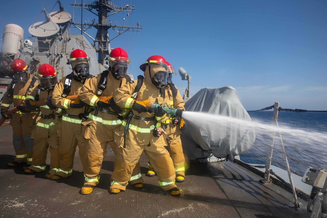 Sailors in firefighting gear stand in line on a ship holding a hose which is spraying water.