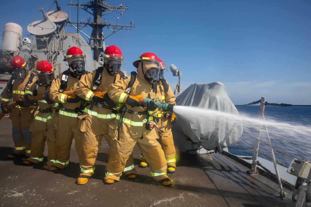 Sailors in firefighting gear stand in line on a ship holding a hose which is spraying water.