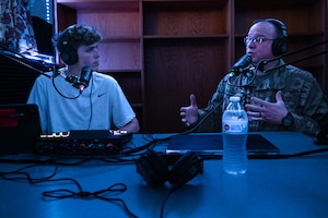 Two men, one in uniform on the right, sitting in front of microphones in a blue-lit studio