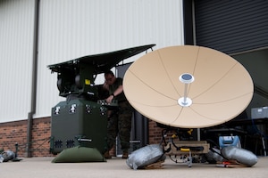 A large, white satellite at right. At left, behind the satellite, a man in a United States Marines Corps uniform adjusting settings on a device located next to the satellite.