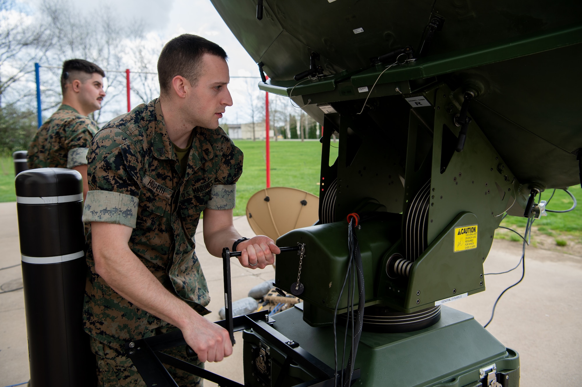 A man in a United States Marine Corps uniform cranks a lever on a satellite to adjust its position. In the background, a man with a mustache with similar uniform. Both men are outside.