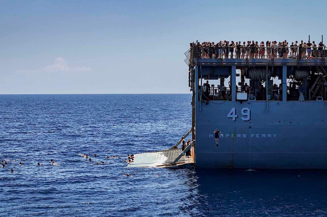 Sailors exit the aft starboard fantail of a Navy ship and swim in open water as others stand atop the vessel to watch.