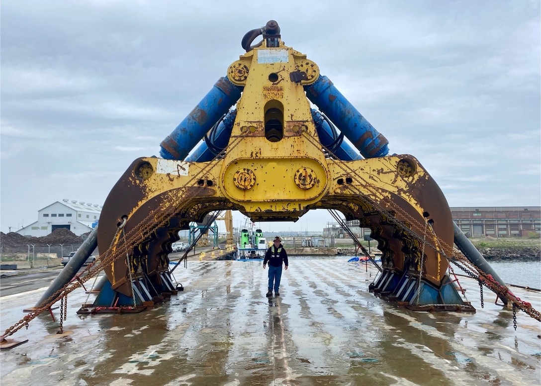 A giant salvage claw is seen sitting on a concrete pad. A man stands beneath the claw, which towers over him, showing the massive scale of the machinery.