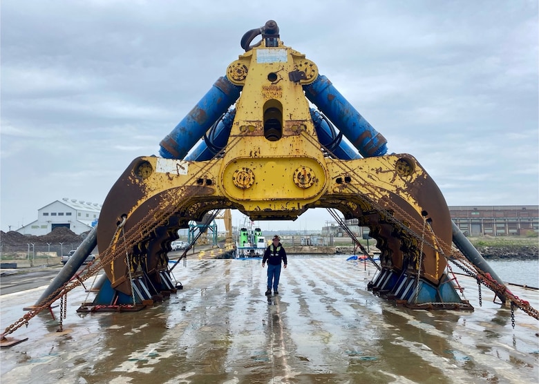 A giant salvage claw is seen sitting on a concrete pad. A man stands beneath the claw, which towers over him, showing the massive scale of the machinery.