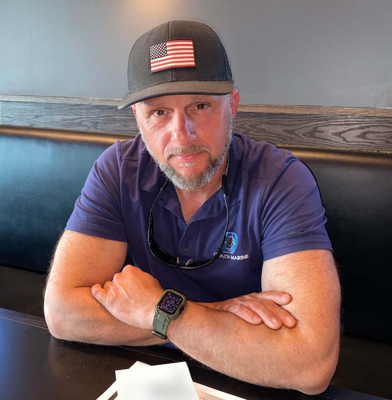 Senior crane operator Vincent DelMaestro sits at a table with his arms crossed. He is wearing a purple shirt and a black baseball cap with an American flag.
