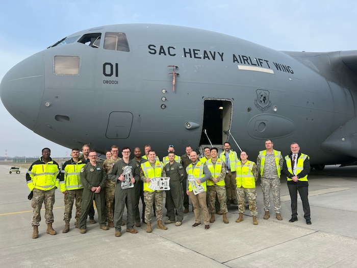 Chief of Staff of the Air Force Strategic Studies Group, Air Mobility Command A4T , Tesseract, Bunker Supply, Boeing and Heavy Airlift Wing personnel pose with the K-Wedge prototype post-test in front of a C-17 Globemaster III at Papa Air Base, Hungary, Jan. 29,2024. The K-Wedge is an installable C-17 loading aid and low-cost solution that eliminates preventable damage during cargo loading operations. (U.S. Air Force photo by 1st Lt. In Bok Lee)