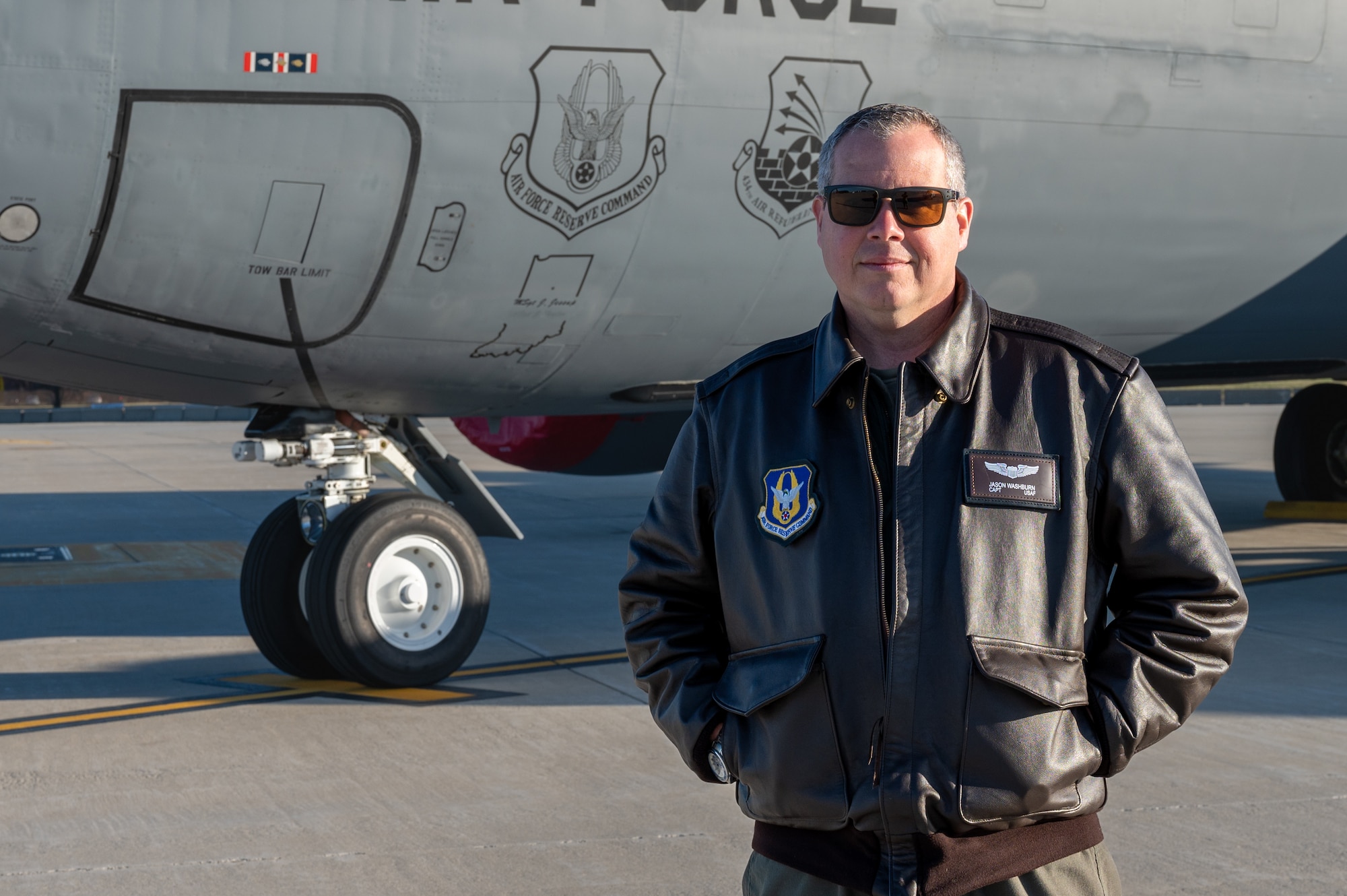 A man in a leather jacket and sunglasses poses for a photo in front of a plane. The plane has two patch decals visible, one for Air Force Reserve Command and the other for the 434th Air Refueling Wing.