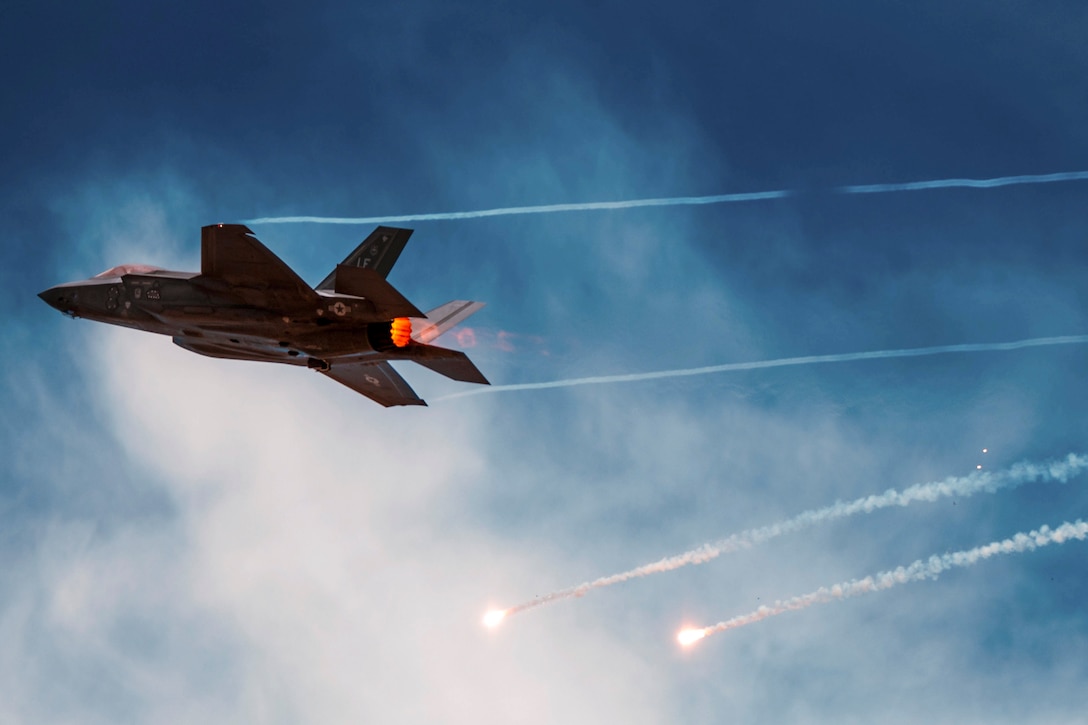 An aircraft leaves streaks while flying in a blue, cloudy sky with two flares descending below it.