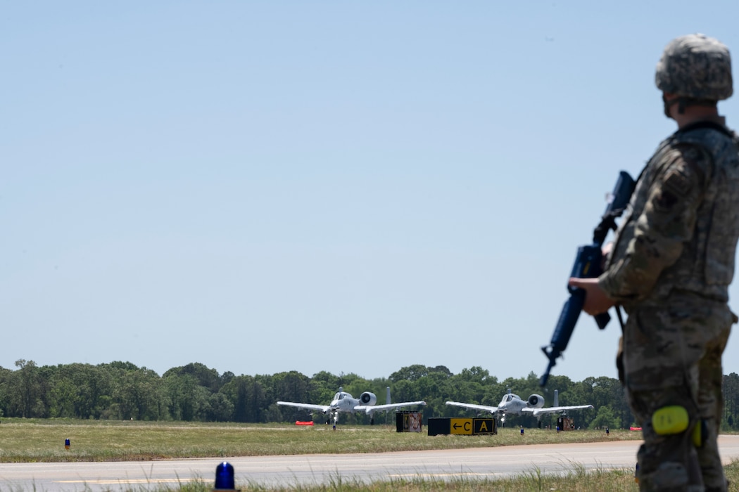 Person standing in fore ground holding security while two A-10C Thunderbolt II's are in the background about to take off.