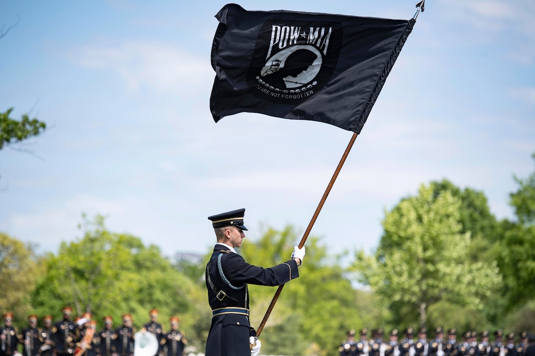 A soldier holds a flag as fellow soldiers in ceremonial uniforms stand in formation in front of trees in the blurred background.