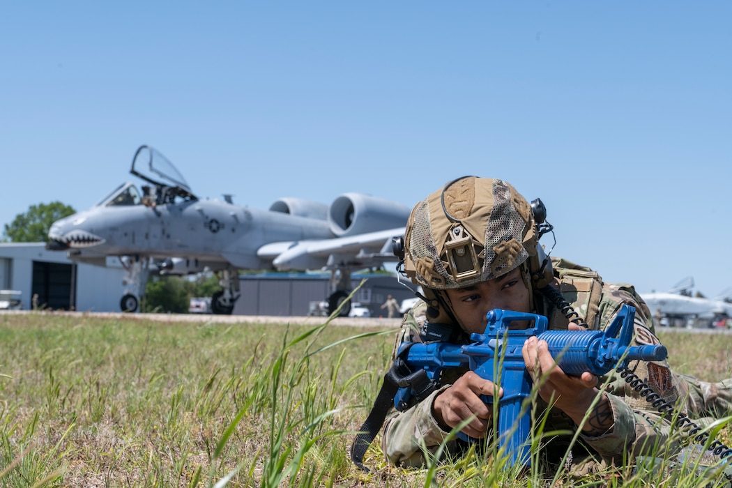 Person laying in grass in the foreground with a dummy weapon, and an A-10C Thunderbolt II in the back ground.