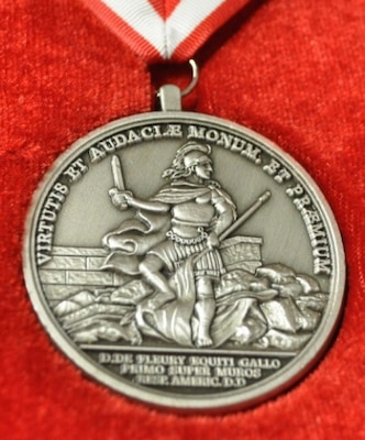 On the medal’s front is “A Memorial and Reward for Courage and Boldness” in Latin. In the center appears the image of a helmeted soldier standing amidst the ruins of a fort, holding in his right hand an unsheathed sword, and in his left the staff of the enemy’s flag, which he tramples underfoot.