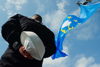 A sailor is seen from behind and below holding a cap while standing at attention. A large, light blue flag with gold stars flutters above the sailor.