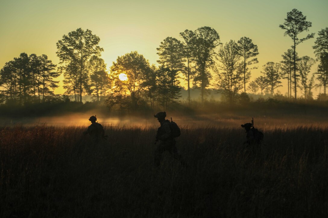 Three soldiers walk through a misty field with the sun behind a row of trees in the background.