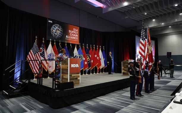 A dark skinned woman in a multicolored dress in front of a multi-flag backdrop stands on stage and sings the National Anthem in a large ballroom. A Marine Corps color guard stands before her.