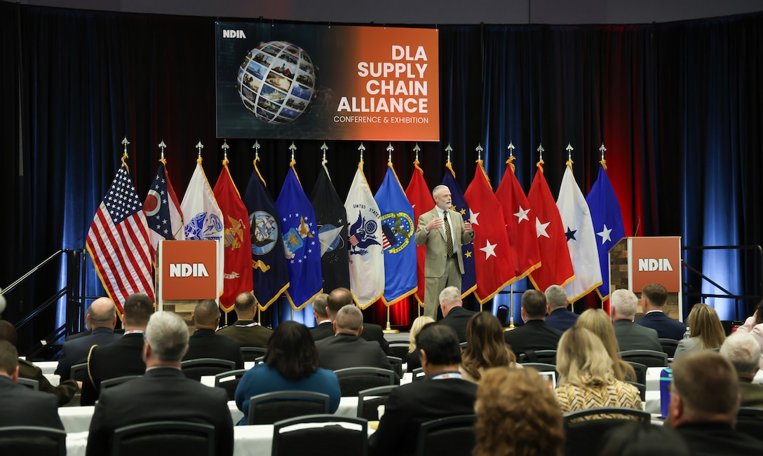 A man in a tan suit in front of a multi-flag backdrop stands on stage and speaks in a large ballroom.