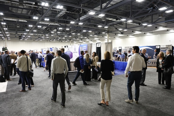 Many people in business attire wander around a large exhibition hall filled with booths.