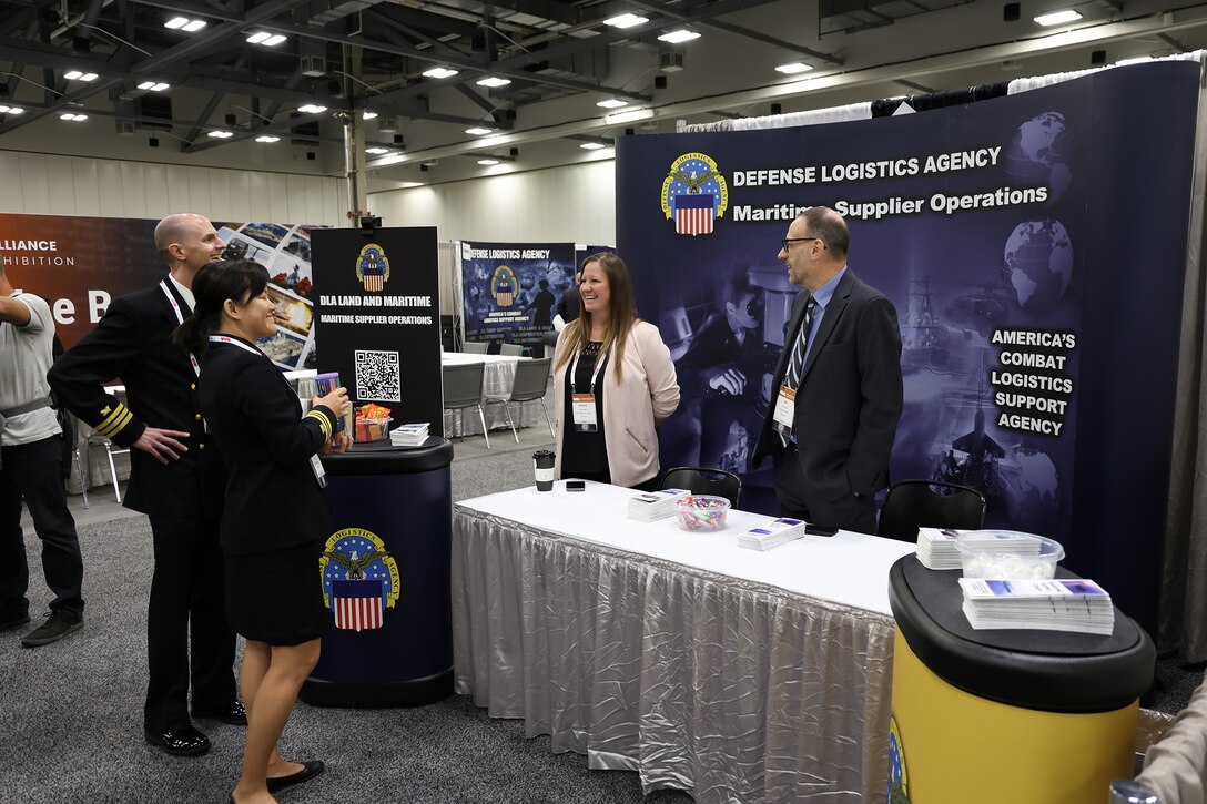 Two Navy personnel, a woman and a man, visit a DLA booth with another man and woman staffing it in a exhibition hall.