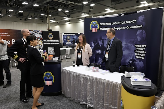 Two Navy personnel, a woman and a man, visit a DLA booth with another man and woman staffing it in a exhibition hall.