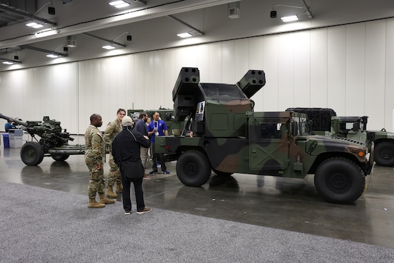 A group of people in business attire, men and women, at a static  display of military vehicles in a exhibition hall.