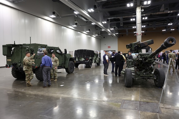 A group of people in business attire, men and women, at a static  display of military vehicles in a exhibition hall.