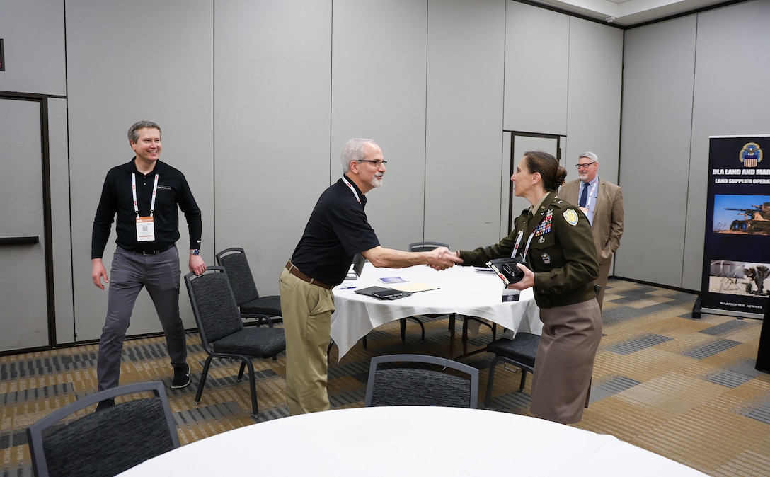 Several people in business suits greet each other in a conference room that is neutral in tone with tables that are round and adorned with white tablecloths. A man meets a woman in a Army uniform in the foreground.