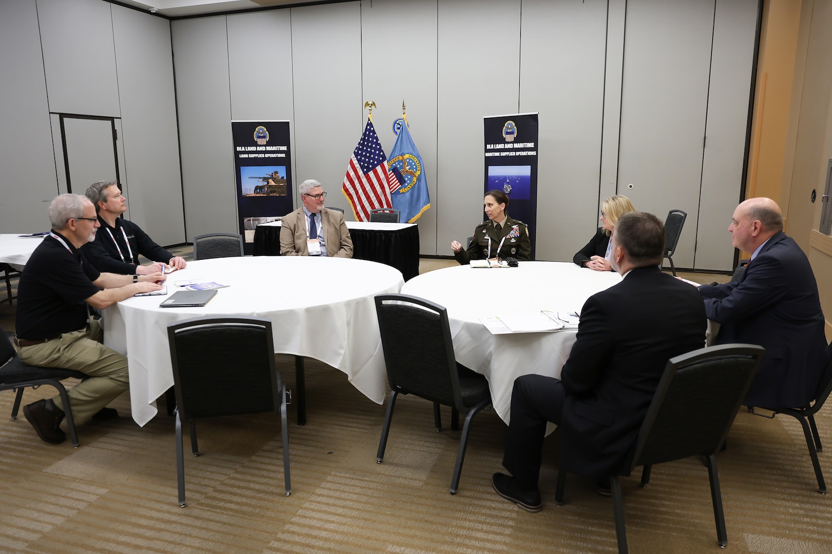 Several people in business suits greet each other in a conference room that is neutral in tone with tables that are round and adorned with white tablecloths.