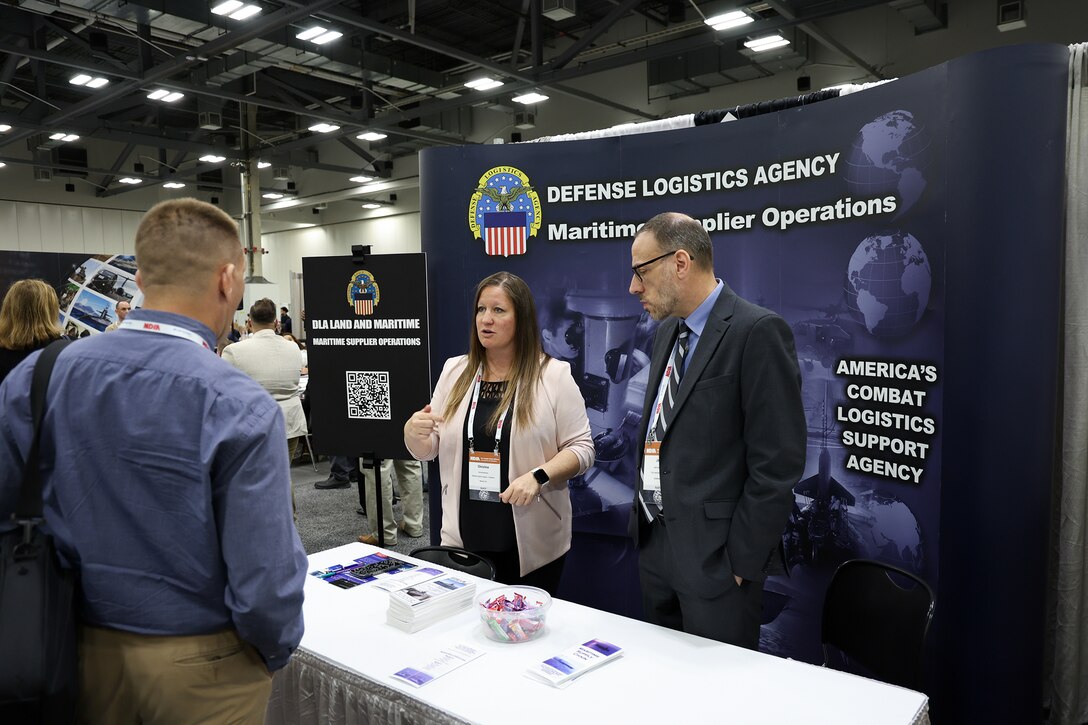 A man visits a booth at the exhibition hall. it is staffed by a man and a woman in business attire. There is a dark backdrop with DLA writing on it.