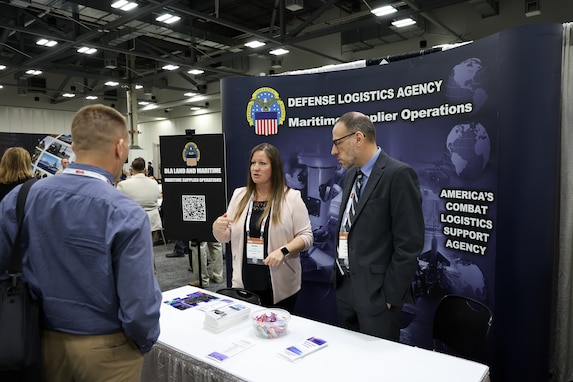 A man visits a booth at the exhibition hall. it is staffed by a man and a woman in business attire. There is a dark backdrop with DLA writing on it.