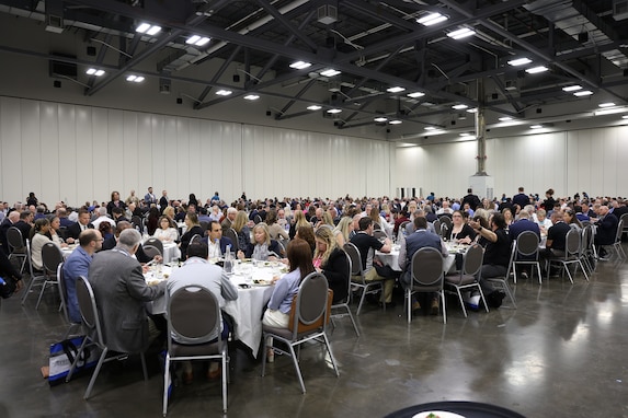 A large group of people in business attire sit down to lunch in a large exhibit hall.