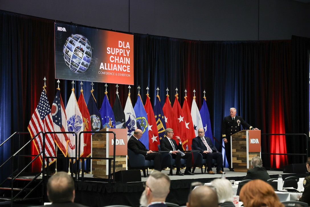 A group of people sit on stage with flags in the background in a large dark ballroom. They are wearing business attire and a gray haired man in a Navy dress uniform stands at the p[odium.