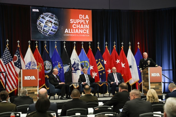 A group of people sit on stage with flags in the background in a large dark ballroom. They are wearing business attire and a gray haired man in a Navy dress uniform stands at the p[odium.