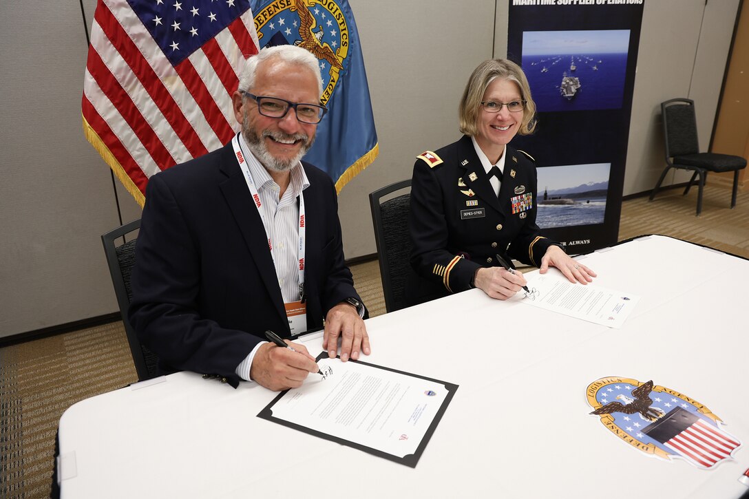 A man in business attire sits next to a light skinned woman in a Army dress uniform in a tan room at a table with a white tablecloth with a flag display behind them.