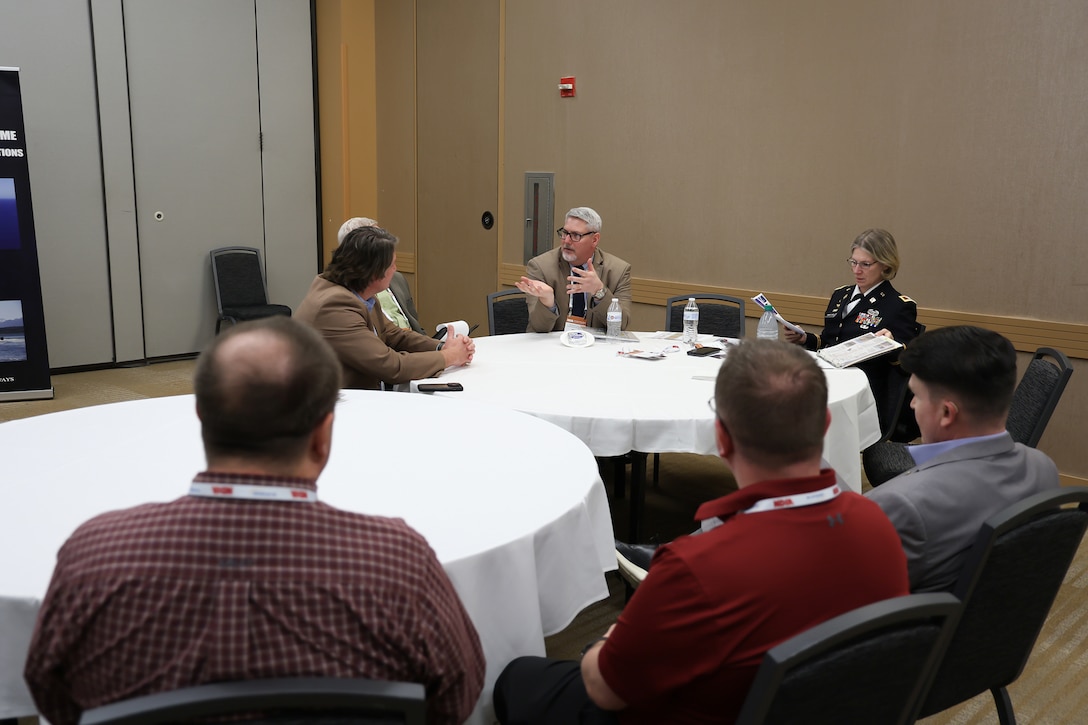 A group of people in business dress chat in a tan room with round white tables.