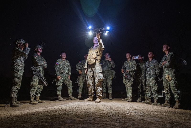 Service members surround and watch as a Marine launches a small drone with lights at night.