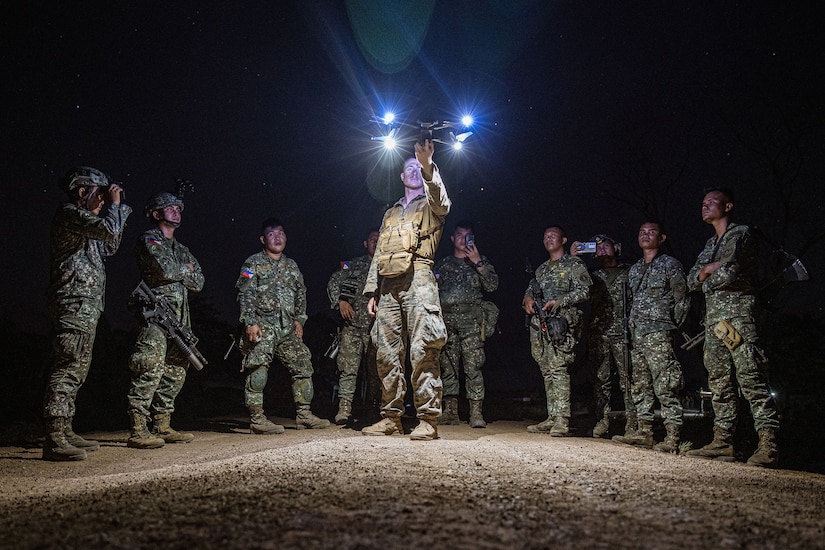Service members watch as a Marine launches a small drone with lights at night.