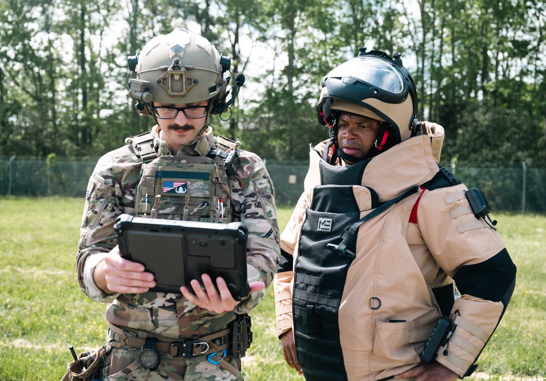 Two Airman, one in a bomb suit and a uniform look at an ipad outside